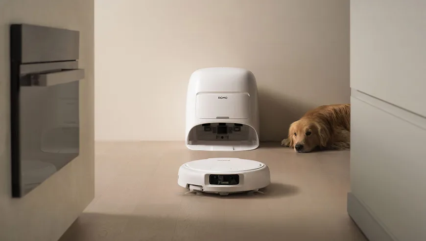 Robot sprzątający ROMO przed stacją dokującą, a w tle pies leży przy ścianie