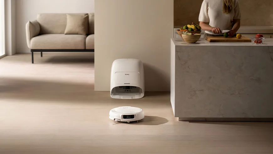 Robot sprzątający ROMO wyjeżdża ze stacji dokującej, a w tle kobieta kroi warzywa w kuchni