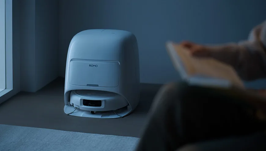 Robot sprzątający ROMO w stacji dokującej o zmroku, obok osoba czytająca książkę