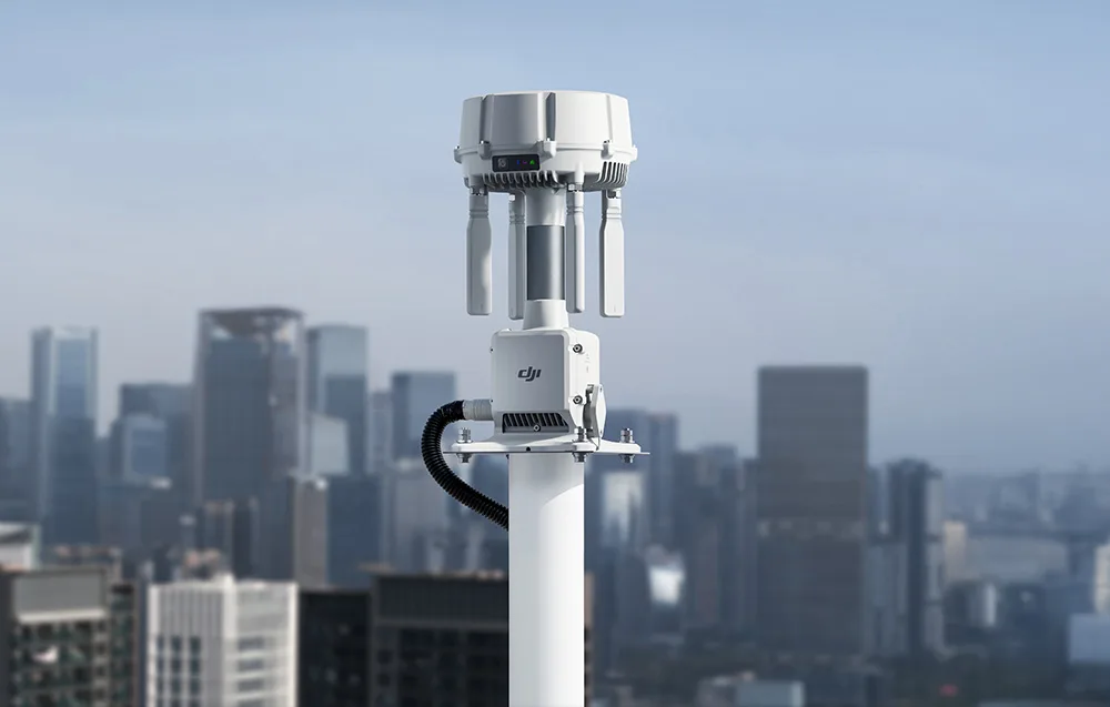 Stacja naziemna DJI z antenami zamontowane na białym maszcie, z rozmazaną panoramą wieżowców w tle