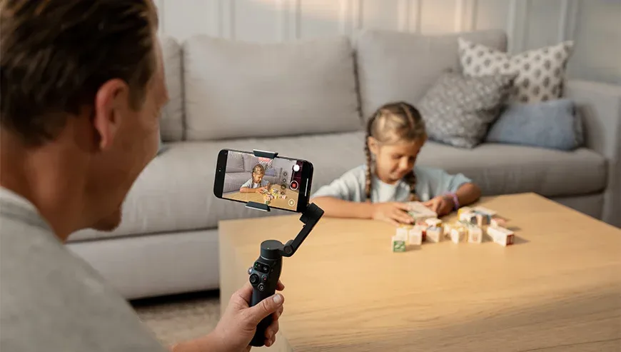 Ojciec nagrywający córkę bawiącą się klockami w salonie za pomocą smartfona na gimbalu DJI Osmo Mobile 8